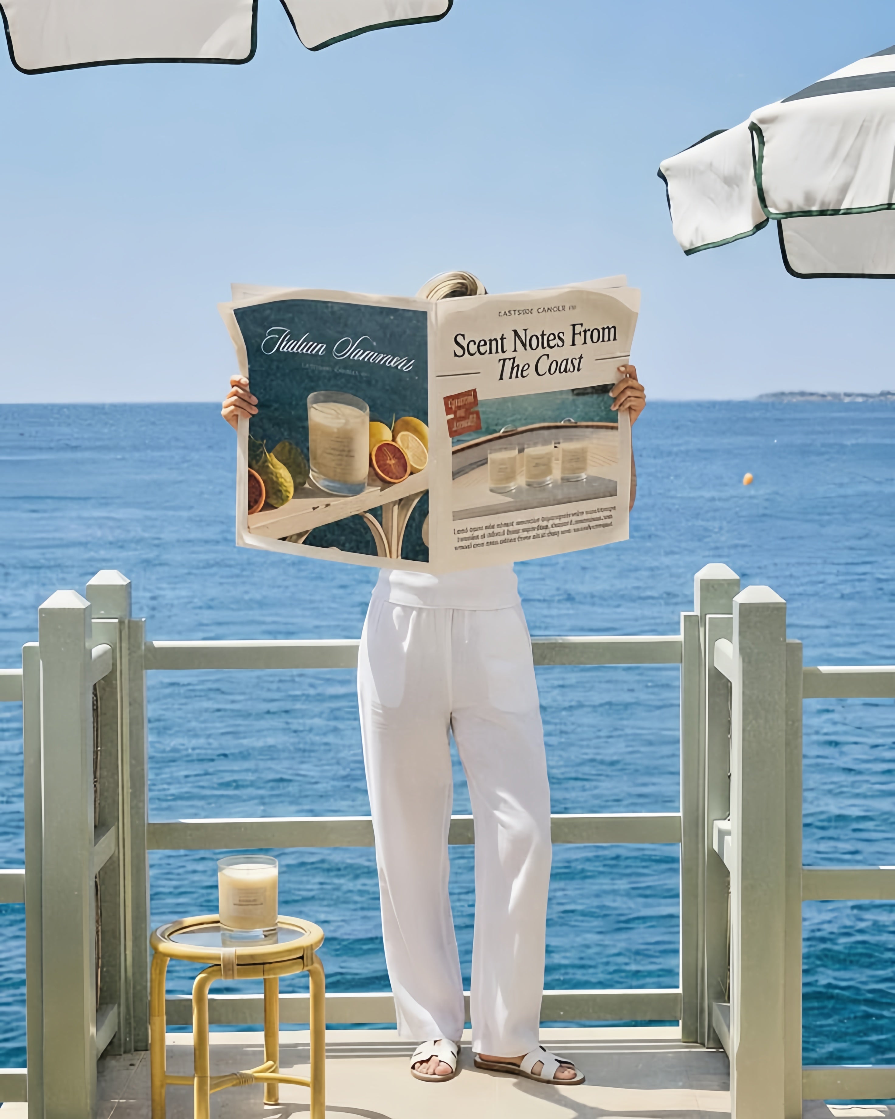 A person stands on a seaside balcony holding a newspaper in front of their face, wearing white pants and sandals. The ocean, blue sky, and patio furniture are visible in the background.